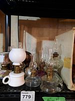 Seven miniature oil lamps arranged on a shelf, including porcelain and glass types with brass burners and glass chimneys.