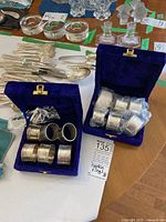 Two blue velvet boxes shown open with silver napkin rings inside; one box contains six rings, the other contains five rings wrapped in plastic. Various silverware and glass pieces visible around.