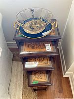 Four stacked wooden nesting tables with glass tops showing detailed carved Asian scenes in light wood, dark wood frames, varying sizes