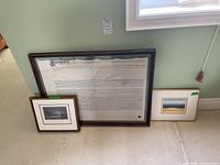 Three framed items leaning against wall under window: large framed document, medium size framed black and white print, and small framed landscape print.