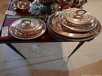 Two large oval silver plated serving dishes with matching lids on a table, showing decorative beaded edges and loop handles on lids.
