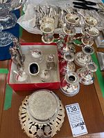 Full lot on table showing all silverplated items including candle holders, condiment jars in a red box, salt cellar, salt and pepper shakers, and trivets.