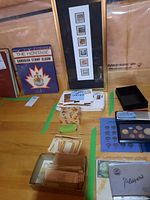 Wide view of framed stamps, stamp albums, loose stamps in envelopes, blue coin holder, and small boxes on wooden table.