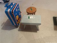 Photo showing a small round wood stool, a rectangular light blue wood stool, and a blue Winnie the Pooh metal suitcase placed on the floor.