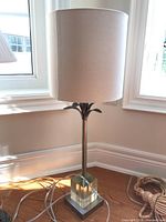 Full view of lamp showing beige cylindrical fabric shade and metal base with lucite accent, positioned near a window.