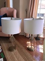 Pair of gold-tone metal table lamps with white drum shades standing on a rug in a room with hardwood floor and pink upholstered chairs.