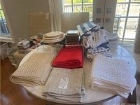 Overview of folded linens including beige knitted textiles, red cloths, beige woven placemats, striped napkins, and white towels arranged on round table.