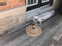 Photo showing the folded light-colored patio umbrella laying horizontally next to the metal stand with a circular lattice base, positioned against a stone wall and wooden deck.