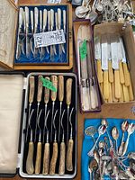 Photo showing boxed cutlery set with Mother of Pearl handles, boxed steak knife set with Bakelite handles, boxed fork set with antler-style handles, and loose spoons/forks on blue cloth.