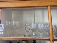 Shelf with assorted stemware glasses including clear Waterford crystal and vintage pressed glass pieces, showing variety of sizes and stem styles.