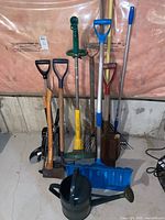 Wide view of assorted yard tools arranged against basement wall: shovels, rakes, axe, watering can, blue shovel in front.