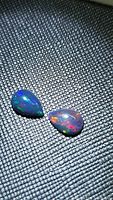 Pair of pear-shaped natural black opals on textured black surface showing multi-color flashes luminously.