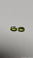 Pair of two natural apple green oval peridot gemstones placed side by side on a light background, showing size and clarity.