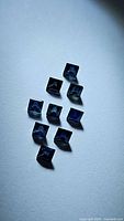 Ten natural princess cut blue sapphires shown on a flat surface with light reflections highlighting the blue color and cut shape.