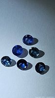 Photo of six natural blue sapphires, round cut, arranged on white surface under lighting showing deep blue color and polished facets.