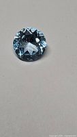 Close-up photo of a single round natural blue topaz gemstone on a gray background showing cut facets and blue color.