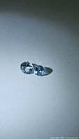 Two round aquamarine gemstones with light blue color on a flat neutral background, showing diamond cuts and clarity.