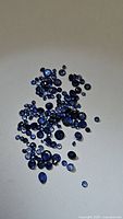 Photo of scattered loose natural blue sapphires on a light background showing variety of sizes and colors ranging from deep royal blue to cornflower blue.