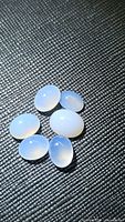 Image of six oval-shaped cabochon chalcedony gemstones on textured black background showing pearly light purple translucency