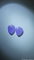 Pair of polished lavender jade gemstones in heart shapes under direct light that highlights their translucent violet color and smooth finish.