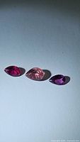 Three pear cut tourmaline gemstones laid out on a flat surface, showing different shades of pink and purple under natural light.