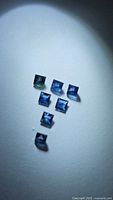 Seven natural, princess cut sapphires with blue-teal hues arranged on a light surface under bright lighting.