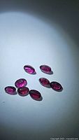Seven oval cut purple pink rhodolite garnet gemstones arranged on a flat surface under bright illumination.