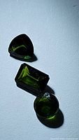 Three natural green chrome diopside gemstones in pear, emerald, and round cuts photographed on a white surface for detail and color display.