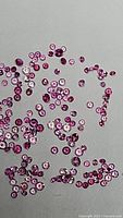 Multiple small round pink sapphires scattered on a neutral gray background, varying in shades of pink and sizes around 1mm to 1.2mm.