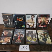 Eight action movie DVDs arranged on a wooden table box featuring titles Gran Torino, Fast & Furious, Mud, Killing Season, Elegy, Gangster Squad, Now You See Me, and Alone in the Dark.