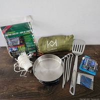 Full lot of camping gear including shelter, skillet, rope, tent spikes, batteries, spatula, and patch material on wooden surface.