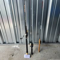 Three fishing rods standing upright, Shakespeare Bank Stik with attached reel, Darwa Eliminator with attached reel, Berkley Enforcer with cork handle and no reel.