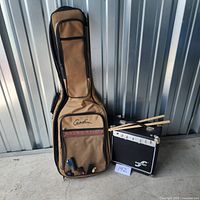 Full view of Godin guitar case standing upright and First Act amplifier with drumsticks on top, all placed on concrete floor against metal wall.