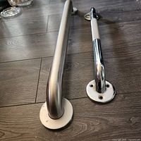 Two shower safety grab bars, one with brushed stainless steel finish and the other with polished chrome finish, shown side by side on a wooden floor.