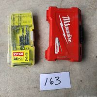 Two drill bit sets in respective cases on concrete floor; yellow RYOBI 35-piece set with transparent lid and red Milwaukee set with logo