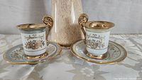 Pair of Royal Vienna demitasse porcelain teacups and saucers placed on a floral tablecloth with a vase in the background.