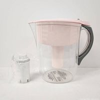Pink Brita water filter pitcher with replacement filter cartridge wrapped in plastic placed side by side on white surface