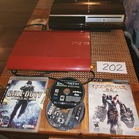 Photo showing two PS3 consoles, one red and one black, with three game discs and cases on a table, and a handwritten number 202.