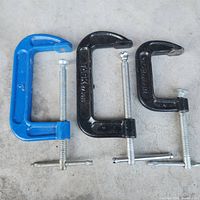 Three metal C-clamps set on a rough concrete floor including one large blue clamp and two smaller black clamps.
