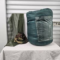 Sleeping bag rolled up, camouflage cap, and army laundry bag displayed on table