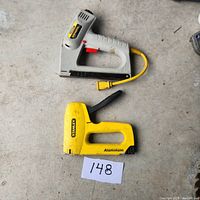 View of two Stanley staplers laid flat on concrete surface, electric stapler with attached yellow power cord and manual aluminum stapler.
