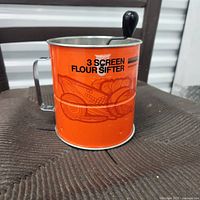 Orange metal flour sifter showing front with '3 Screen Flour Sifter' and bread graphic.