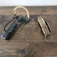 Photo showing vintage black handled slingshot with beige rubber bands and vintage multi-tool utility cutter with wooden textured handle
