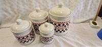 Four Coca-Cola ceramic canisters with checkered red, white, and black designs and lids with round knobs.