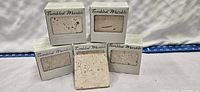 Five 4x4 inch tumbled marble tiles in original packaging displayed with one tile out showing texture and color.