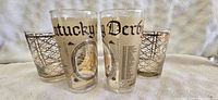 Four highball glasses displayed together on a white textured surface, two official Kentucky Derby 100th Anniversary glasses with gold and black detailing, and two Tostestler gold geometric lattice patterned glasses.