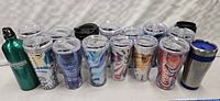 16 Coca-Cola themed tumblers and water bottles arranged in rows on a white surface. Various colorful cruise designs on plastic tumblers with clear lids and straws, plus three metal bottles/tumblers.
