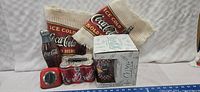Showcases the Coca-Cola glass set in white box with retro logo, two cream and red woven Coca-Cola tablecloths with fringe edges, the bottle-shaped timer, and the six-pack lunch box partially visible.