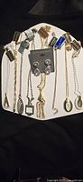Front view of 8 necklaces and one set of Clip Earrings attached to a display card, showing various gold-tone chains and different pendant shapes including geometric and decorative designs.