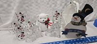 View of all items on white cloth background including six snowman mugs, two teddy bear candle holders, white teddy bear figurine, and snowman card holder
